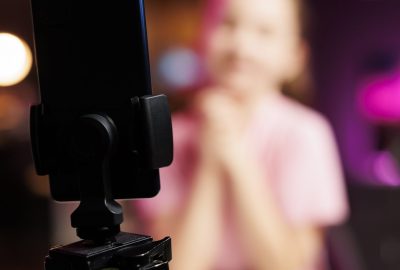 Close up of phone on tripod stabilizer used by child sponsored by brand to showcase message on children channel. Focus on smartphone recording kid in blurry background doing influencer marketing